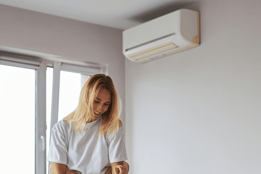 A smiling young woman in a white t-shirt enjoying the cool air from a wall-mounted air conditioner in a light-colored room.